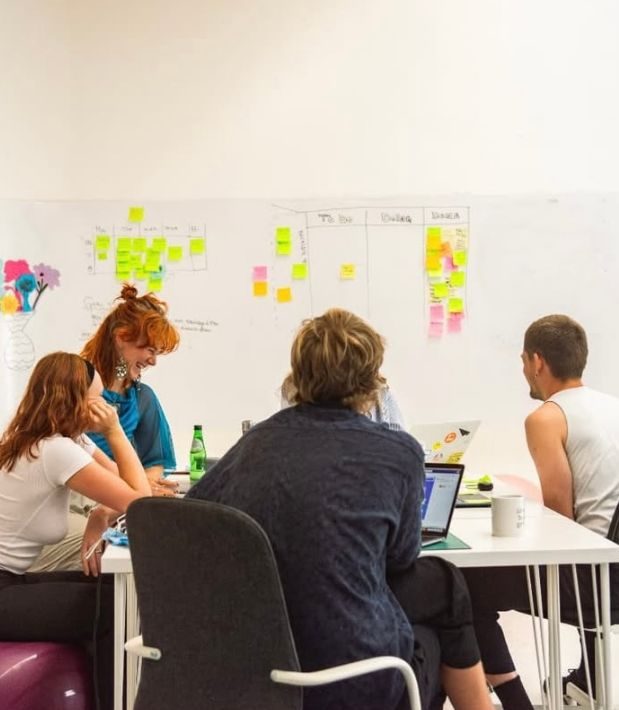 Equipe reunida ao redor de uma mesa, planejando com post-its em um quadro branco.