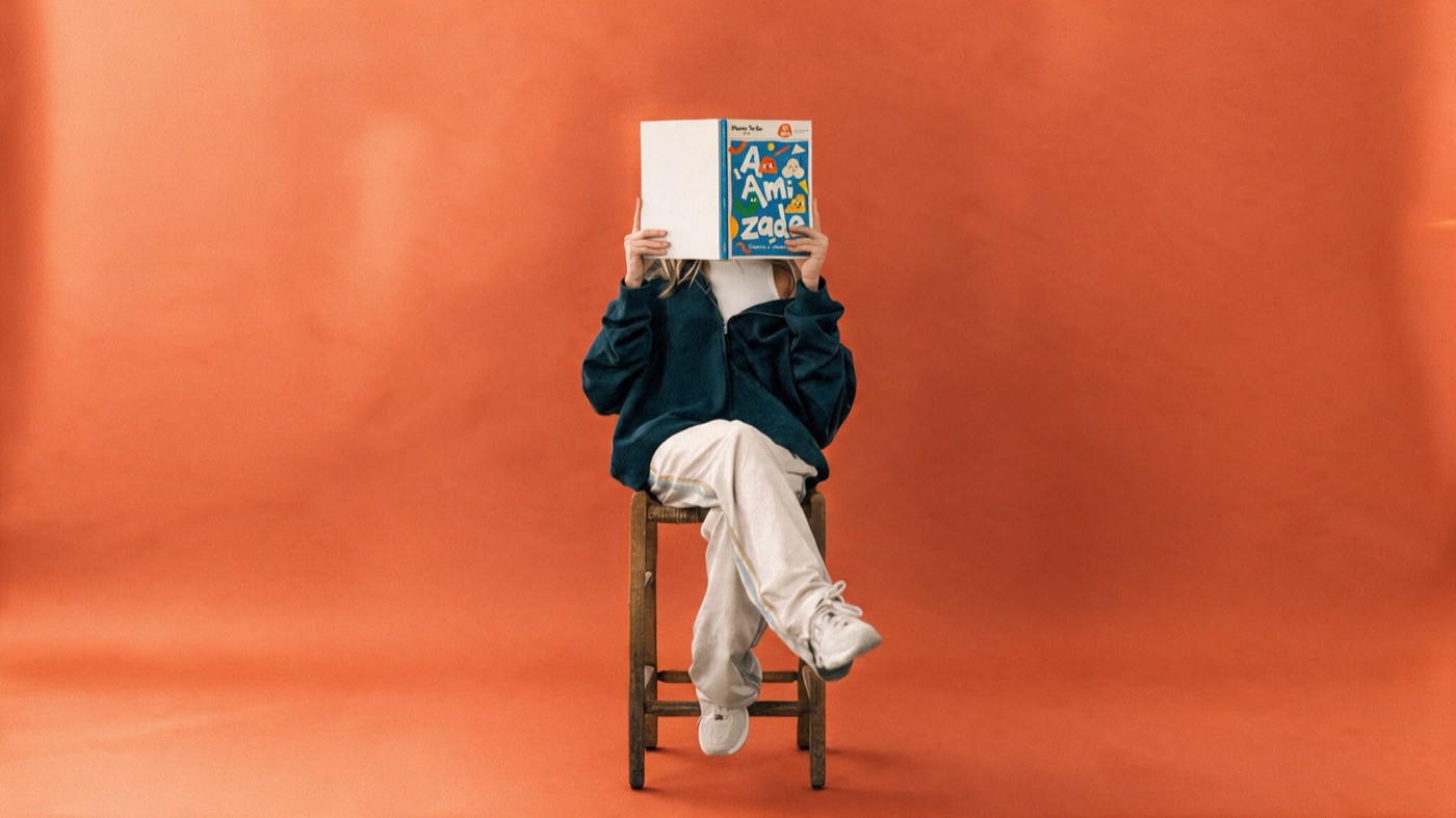 Estudante sentada num banco de madeira lendo o livro A Amizade, da Plann To Go Life, diante de um fundo laranja.
